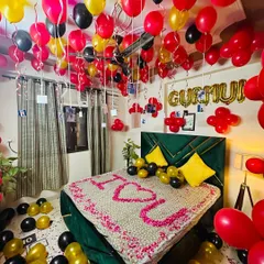 A romantic bedroom setup with red, black, and gold balloons. The bed is covered in petals spelling "I <3 U", and balloons with photos hang from the ceiling.