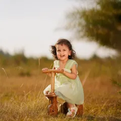 A happy toddler laughs while sitting on a wooden tricycle in a field of tall grass, bathed in beautiful golden hour light.