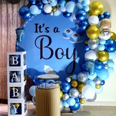 A classic 'It's a Boy' baby shower decoration. The setup includes a large circular backdrop, a blue, white, and silver balloon garland, and 'BABY' blocks.