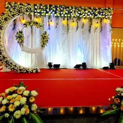 A wide view of a naming ceremony stage inside a hall. The floral moon arch is complemented by elegant white curtains and warm lighting, creating a serene and beautiful ambiance for the event.