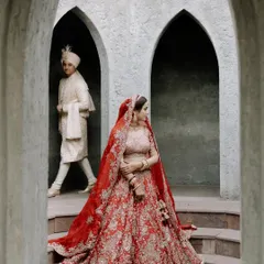A moment of quiet anticipation. The bride, Priya, in her stunning red lehenga, stands framed by ancient arches as her groom, Nishant, walks in the background. A story in a single frame.