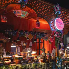 A vibrant and colourful food stall setup for a festive event. The decor, with its traditional umbrellas and floral garlands, complements the rich Indian cuisine being served.