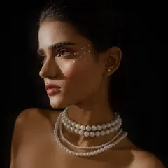 A classic profile beauty shot against a dark background. The lighting carves out the model's features and highlights the layered pearl necklace and eye makeup.