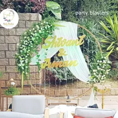 A personalized circular backdrop for an anniversary or engagement party. The couple's names in gold are set against a white drape, framed by an arch of fresh white flowers and greenery for a romantic, garden-party feel.