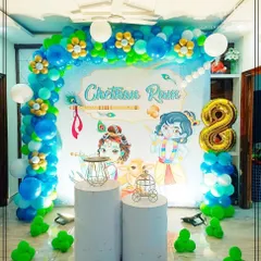 A divine Little Krishna theme for a birthday celebration. This backdrop features adorable illustrations of Krishna and Balaram, a peacock feather motif, and a blue and green balloon arch decorated with floral elements.
