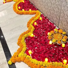 A traditional floor decoration (rangoli) made with flower petals for a housewarming event. This design uses vibrant yellow marigolds and red rose petals, outlined with small electric candles for a warm glow.