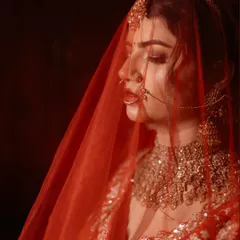 A close-up bridal portrait, with the sheer red veil creating a soft, dreamy effect. This image captures the bride's delicate features and the rich color of her wedding lehenga.