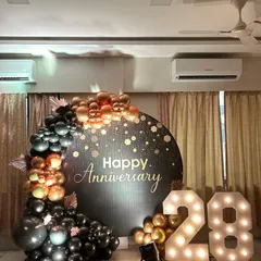 A chic 28th anniversary decoration for a home celebration. This setup features a stylish black backdrop with gold confetti details, a black and gold balloon garland, and large light-up numbers.