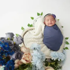 This smiling newborn is swaddled in a deep blue wrap, resting in a heart-shaped basket adorned with flowers. We love capturing those fleeting, happy little moments.