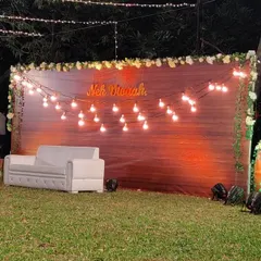 For a warm and intimate feel, we designed this rustic engagement backdrop. It features a rich wooden panel, a personalized neon sign with the couple's name, and strings of cozy Edison bulbs, perfect for an outdoor lawn party.