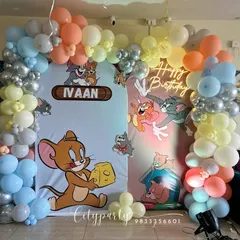 A classic Tom and Jerry theme for a 5th birthday. This setup features a dual backdrop with the iconic characters, a pastel balloon arch, a 'Happy Birthday' neon sign, and a number '5' balloon column.
