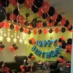 A simple yet festive birthday room decoration. The ceiling is covered with red and black balloons with hanging ribbons, and "Happy Birthday" foil balloons are placed on the wall to create a party vibe at home.