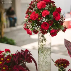 A tall, elegant centerpiece featuring a vibrant mix of red carnations and roses in a clear glass vase. This style adds height and drama to a tablescape without obstructing the view, making it ideal for gala dinners and award ceremonies.