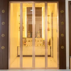 A serene pooja room with calming, gold-tinted glass doors that fold away. The interior is well-lit, creating a peaceful and divine atmosphere.