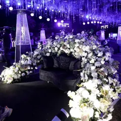 A dramatic and luxurious seating area for a winter reception. A black velvet sofa is enveloped in a cascade of white flowers, set against a backdrop of hanging crystal strings and purple uplighting.