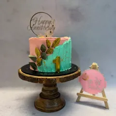 This anniversary cake features a contemporary two-tone design with pink and teal buttercream. A spray of golden leaves climbs the side, tying in with the gold '1' and the elegant 'Happy Anniversary' topper.