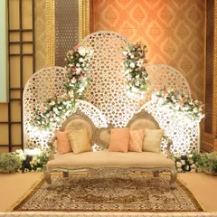 An elegant stage setup with a vintage-style sofa, set against a white jaali-work backdrop adorned with delicate pink and white floral arrangements.