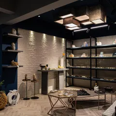 This corner of the boutique features custom-made, backlit stone veneer cubes that appear to float from the ceiling, creating a stunning light installation. Below, minimalist metal shelving provides a clean, industrial contrast to the textured brick wall.