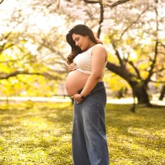 A casual and beautiful outdoor maternity portrait. The expecting mother cradles her bare bump against a backdrop of lush, sunlit trees.