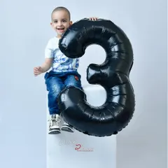 A happy three year old boy celebrating his birthday in the studio. He is sitting on a white block next to a large black balloon in the shape of the number three.
