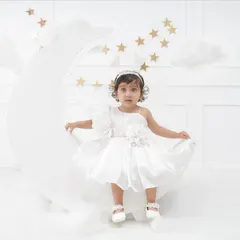 A whimsical 'Moon & Stars' themed portrait of a little girl in a beautiful white dress, sitting on a large crescent moon prop surrounded by clouds and stars.