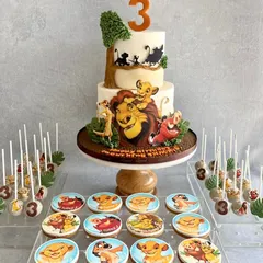 A full dessert table for a Lion King fan! The two-tiered cake features hand-painted scenes from the movie, and it's surrounded by themed cookies and cake pops with all the beloved characters.