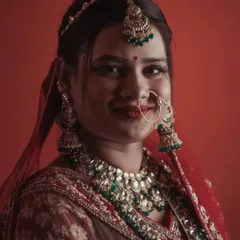 A classic bridal portrait against a simple red backdrop. The direct, gentle smile of the bride showcases her happiness, while the lighting beautifully illuminates her green and red kundan jewelry and traditional attire.