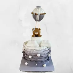 A whimsical two-tiered cake featuring a cute teddy bear in a hot air balloon. The grey ombre ruffles and pearl details make this perfect for a dreamy baby shower.