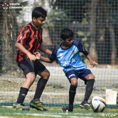 An intense one on one battle on the turf, as our player in the red and black kit shields the ball from the opposition.