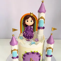 A fairytale castle cake designed for a little princess's fourth birthday. This piece features hand-sculpted turrets with golden flags, a custom princess figurine in a ruffled purple dress, and a rainbow arch for an extra touch of magic.