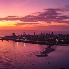 The Mumbai coastline and Marine Drive bathed in the soft, purple and pink hues of a tranquil sunset. This drone shot captures the serene meeting of the city and the Arabian Sea, with the tide creating patterns on the shore.
