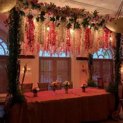 An elegant VIP dining table setup, perfect for hosting key stakeholders or award winners. The table is framed by a beautiful overhead structure with hanging floral arrangements and warm incandescent bulbs, creating an exclusive and intimate setting.