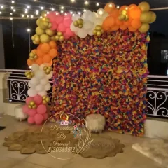 A vibrant and colorful photo backdrop made of multi-colored paper flowers. The wall is framed with an organic balloon garland in pink, orange, and white, creating a cheerful spot for pictures.