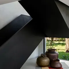 A different angle of the sculptural staircase, emphasizing its sharp, geometric form. The space beneath is adorned with traditional brass pots, creating a dialogue between modern architecture and cultural artifacts.