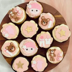 An assortment of pink-themed baby shower cupcakes on a wooden board. The designs include fondant teddy bears, baby clothes, gold stars, and baby faces with knitted hats, demonstrating a cohesive and detailed theme for celebrating a baby girl.