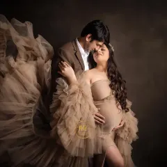 A romantic and dramatic couple's portrait. The mother-to-be, in a stunning beige ruffled gown, shares a close moment with her partner against a dark, moody backdrop.