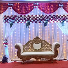 A classic wedding stage design featuring elegant red and white drapes. The backdrop is illuminated by a curtain of fairy lights, complemented by a floral cornice and matching bouquets for a timeless look.