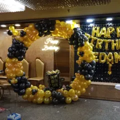 A stylish black and yellow balloon ring setup in a banquet hall. This design includes a 'Happy Birthday' neon sign and a black sequin wall with foil letters spelling out the name.