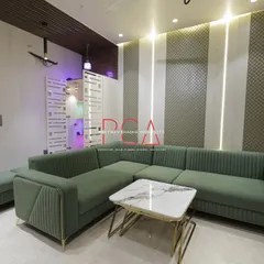 The living room of The Cursive Abode, featuring a plush green sectional sofa, a marble-topped coffee table, and a feature wall with textured panels and integrated vertical lighting. The space is an example of modern comfort and luxury.
