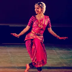 A vibrant stage photograph from a solo Bharatanatyam recital. The bright pink and gold costume, combined with the expressive smile, captures the joyful spirit of the dance.