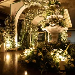 This grand entrance features a stunning fountain surrounded by an abundance of white flowers and greenery. The warm uplighting and arched structures create a luxurious and welcoming atmosphere for an evening event.