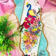 This peacock wall art is set against a soft pink background, with a lush green plant providing a natural setting. The piece features intricately sculpted feathers and flowers, accented with pearls.