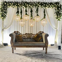 A regal seating arrangement for a Walima or Nikaah. A vintage-style sofa is set against a backdrop of white drapes, with hanging crystal chandeliers adding a touch of opulence.