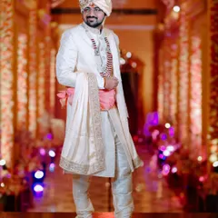 A full-length portrait of the groom in his elegant off-white sherwani. The warm, glowing background of the venue adds a touch of grandeur to his sharp look.