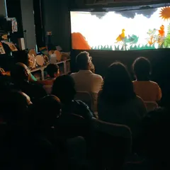 A view from the audience as they watch the Chitrakathi performance. The room is dark, and all eyes are fixed on the illuminated screen where the ancient story of the birds and the hunter unfolds.