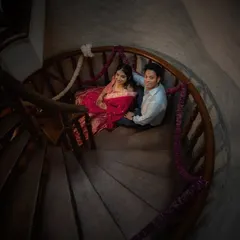 A high-angle shot of a couple on a dark wood spiral staircase. The composition draws the eye down, creating a sense of intimacy and romance within the architectural space.