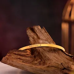 A gold-plated cuff engraved with 'HRITHIK', resting on a piece of driftwood. The warm, rustic setting complements the rich gold tone of the bracelet.