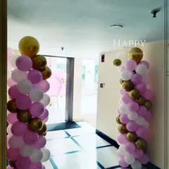 Elegant balloon pillars in pink, white, and gold lining the hallway. These decorative elements guide guests to the main event area while enhancing the party's ambiance.