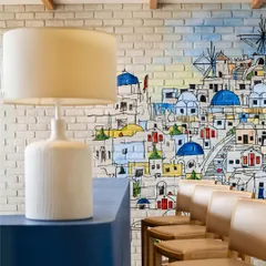 A detail from a cafe space, where a hand-painted mural of a Mediterranean village adds a touch of whimsy and color. The exposed brick wall and simple bar stools create a rustic, charming vibe.