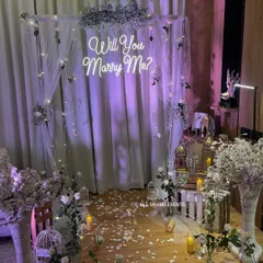 This is how we turn a simple room into a romantic proposal spot. The setup features a 'Will You Marry Me?' neon sign against a backdrop of sheer white drapes and fairy lights. We scattered white rose petals and placed flameless candles to create a warm, intimate, and absolutely magical atmosphere.